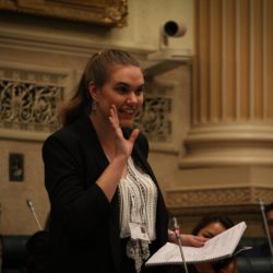 Image of Eloise Duncis standing in the House of Assembly in South Australian Palriament wearing a black suit and white shirt with her right hand raised in the middle of making a statement.